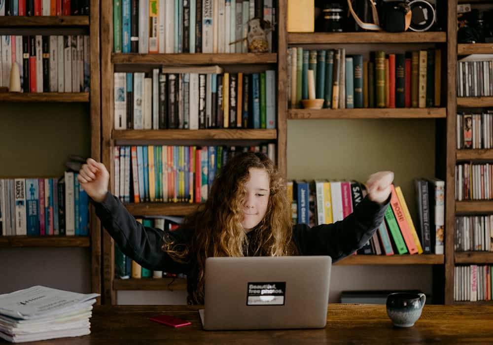 Jovem com braços abertos sentada em uma mesa de cadeira olhando para um notebook. Ao funo, uma estante de madeira repleta de livros.