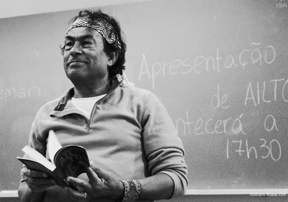 A foto é em preto e branco. Na imagem, Ailton Krenak,, um homem com traços indígenas e de cabelos pretos ondulados, aparece do lado esquerdo da imagem segurando um livro semi-aberto nas mãos. Ele veste um cardigã com a gola aberta e uma faixa estampada na cabeça. No fundo, há uma lousa com palavras escritas em giz branco informando sobre um evento que acontecerá às 17h30.